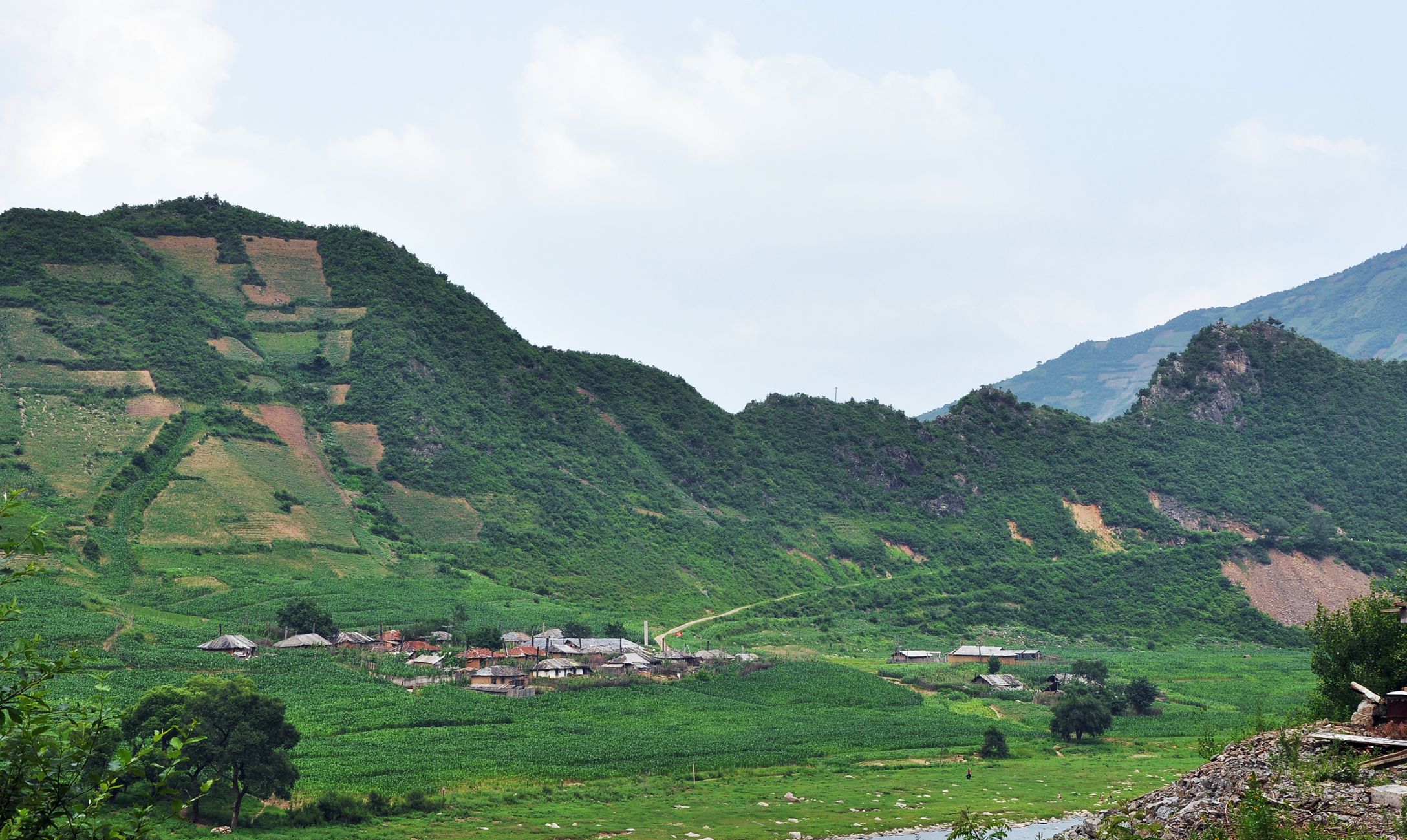 图为朝鲜乡村风貌,一座村庄坐落在山脚下,山脚和山坡上都种满了农作物