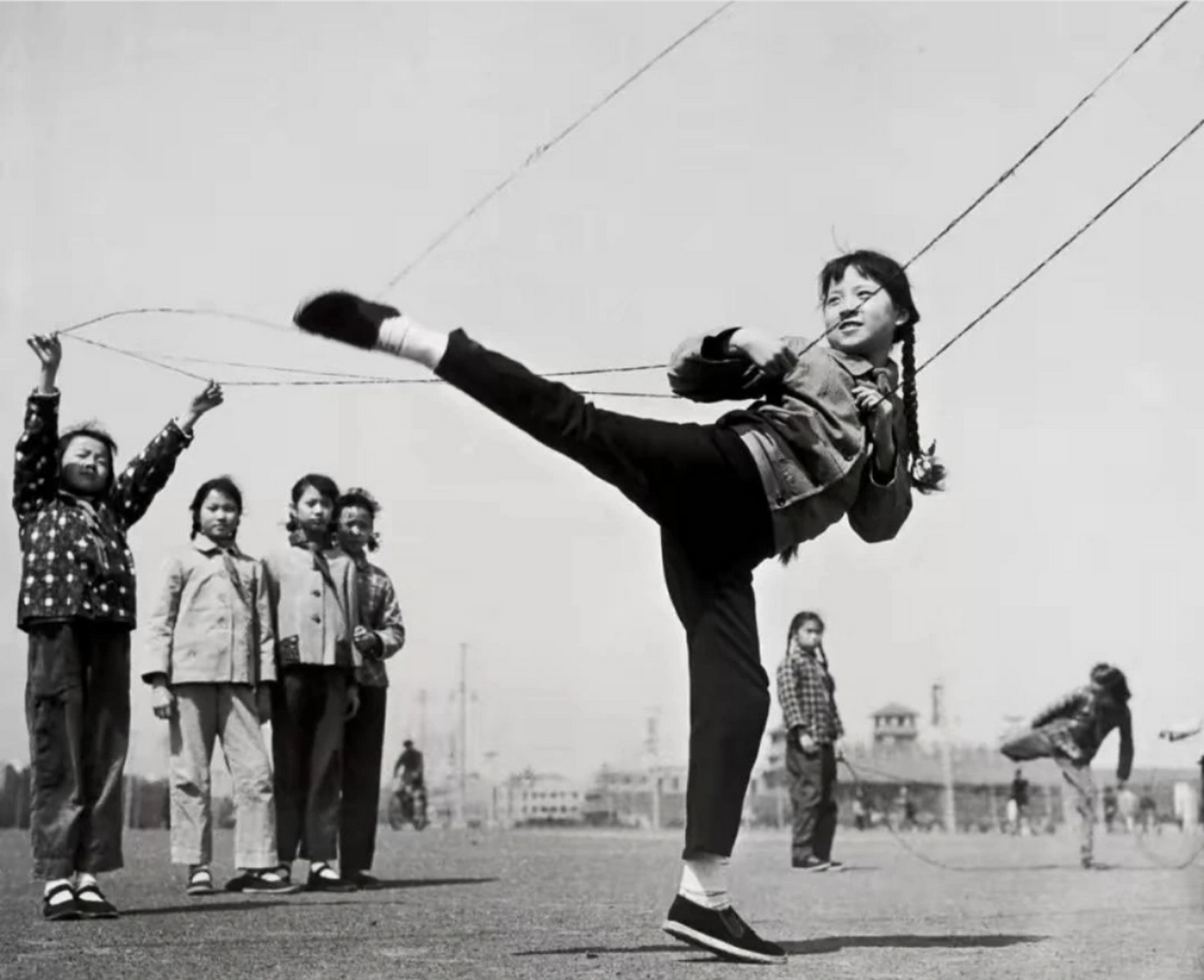 照片中,我们看到一位女同学在操场上跳皮筋,周围是那个时代的建筑物和