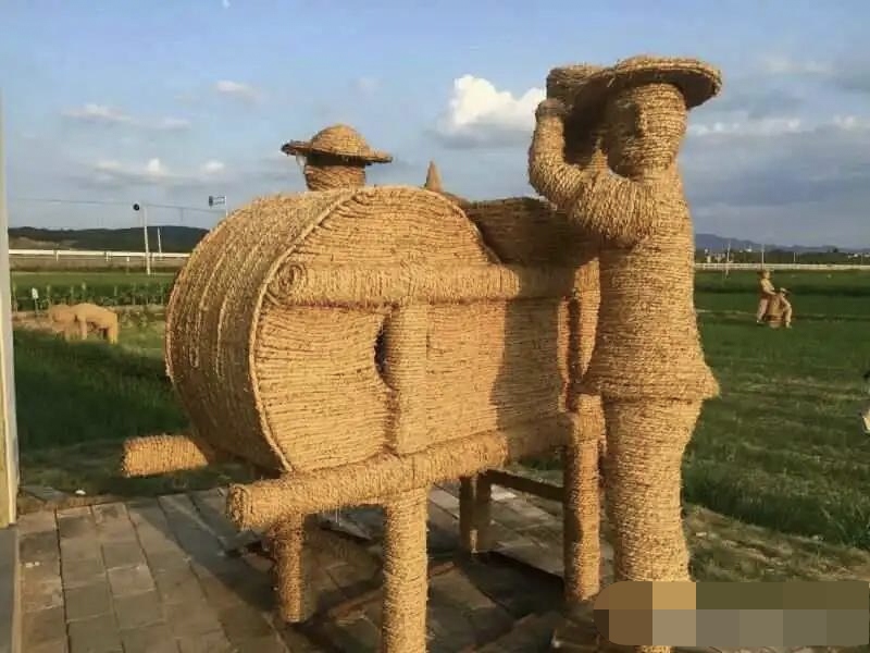 一个大型的稻草雕塑,由稻草编织而成的人形象和农具立在田野之中,展现