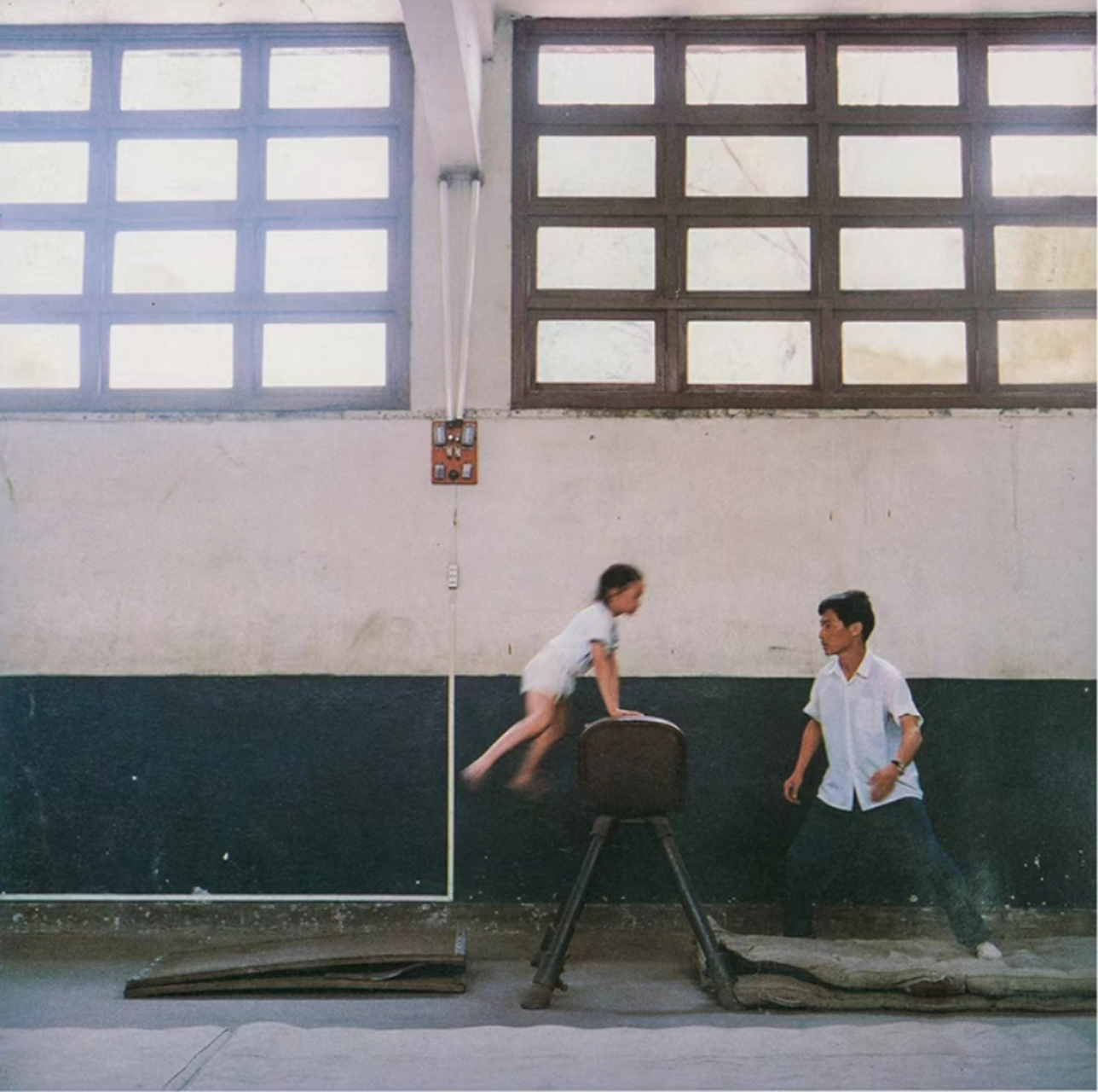 日本摄影师拍摄的八十年代老照片,正在练习跳马的小女孩,教练在旁边接