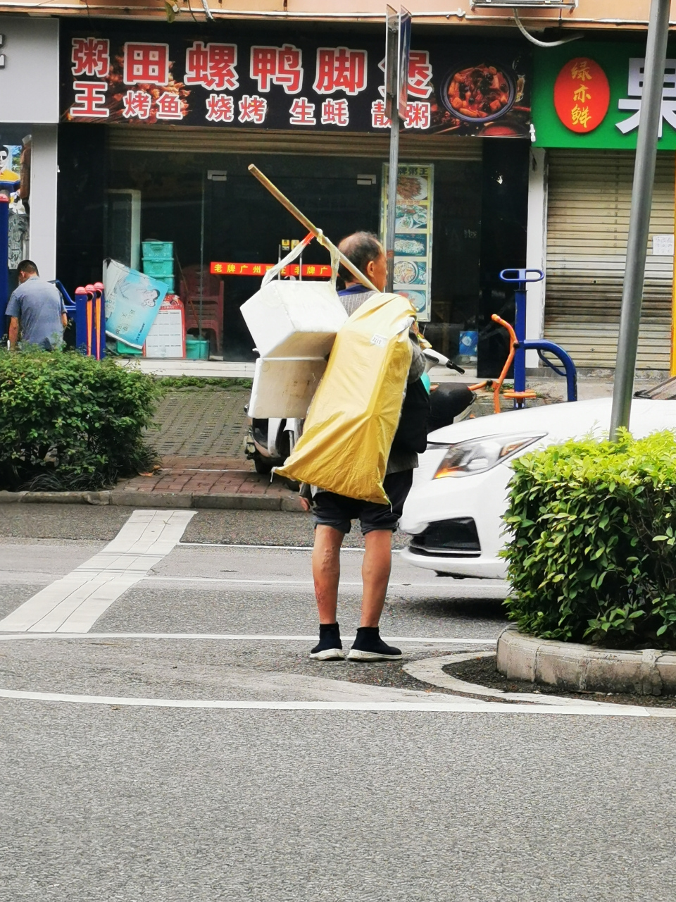 一位老大爷用一根长棍挑着一个黄色的麻袋和一个盒子走在路上,此时