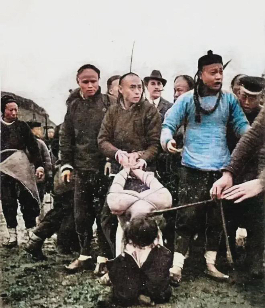 老照片,画面中一名囚犯双手被反绑,麻木的跪在地上,其中一人扯着犯人