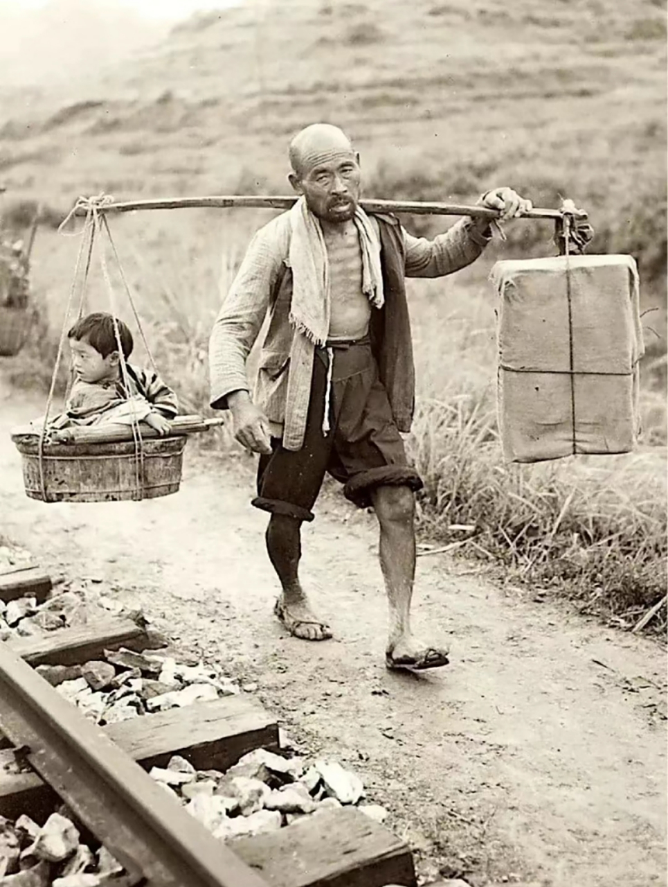 民国老照片,一位老爷爷穿着草鞋,挑着担子从铁轨旁边经过,担子一头挑