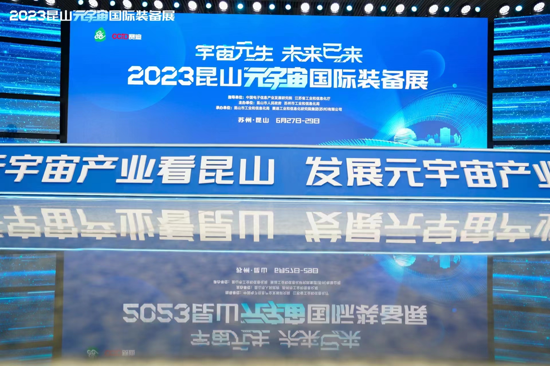 图片展示了一个带有文字的大型显示屏，前方有模糊的文字反射，背景为蓝色调，可能是某个会议或活动的现场。