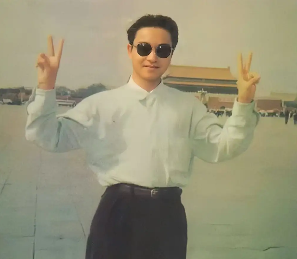 照片中张国荣穿着黑西裤和白衬衫,戴着黑色墨镜,双手比剪刀手,帅气之