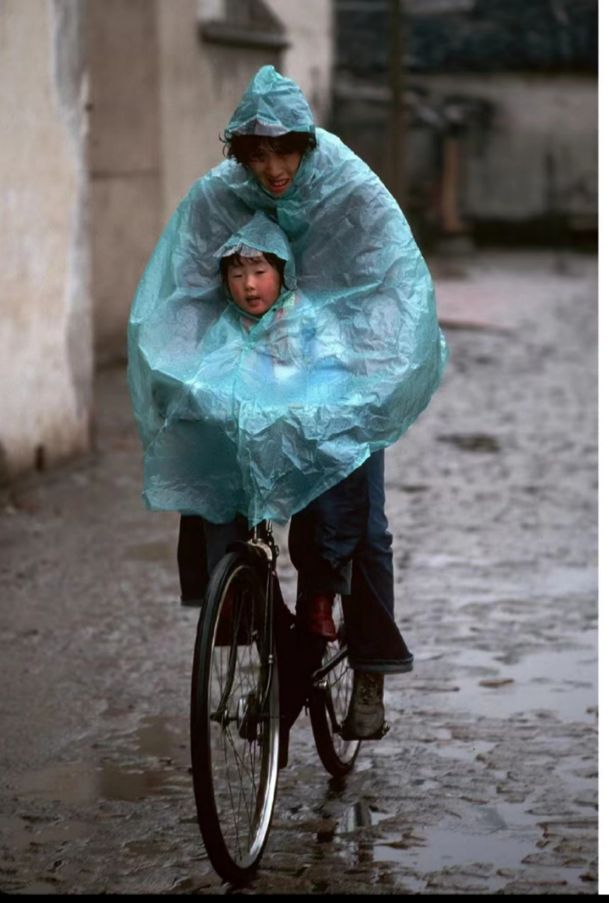 苏州,在雨中,一位母亲穿着雨衣骑着自行车驮着孩子,在巷道里前行!