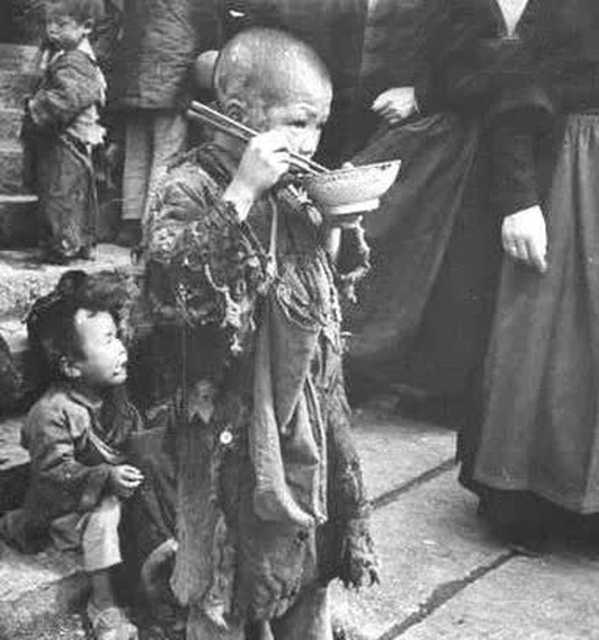 图中的小男孩也是如此,他衣衫褴褛,光着脚,端着碗充饥地站在