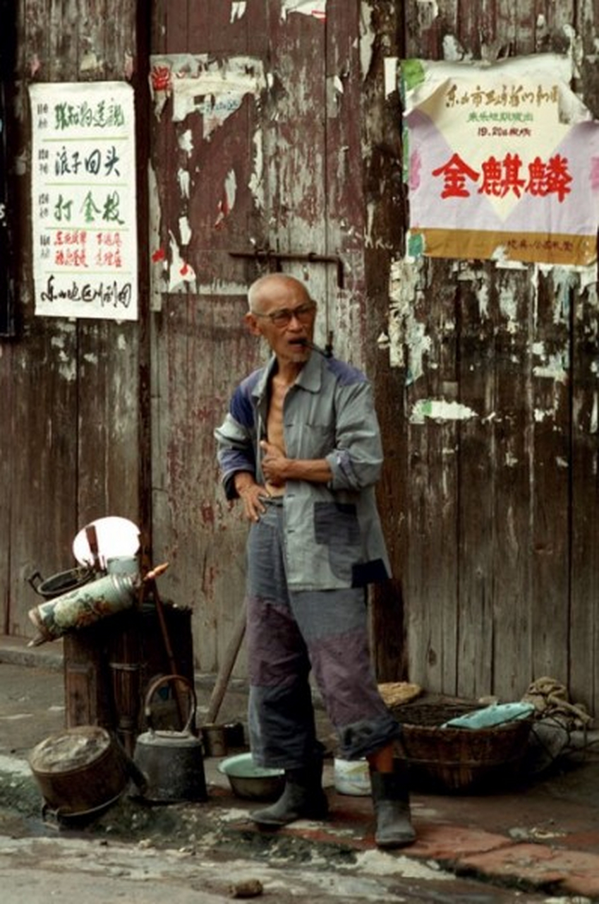 一个老大爷穿着补满了补丁的衣服,咬着烟斗站在街边上,旁边放着一些