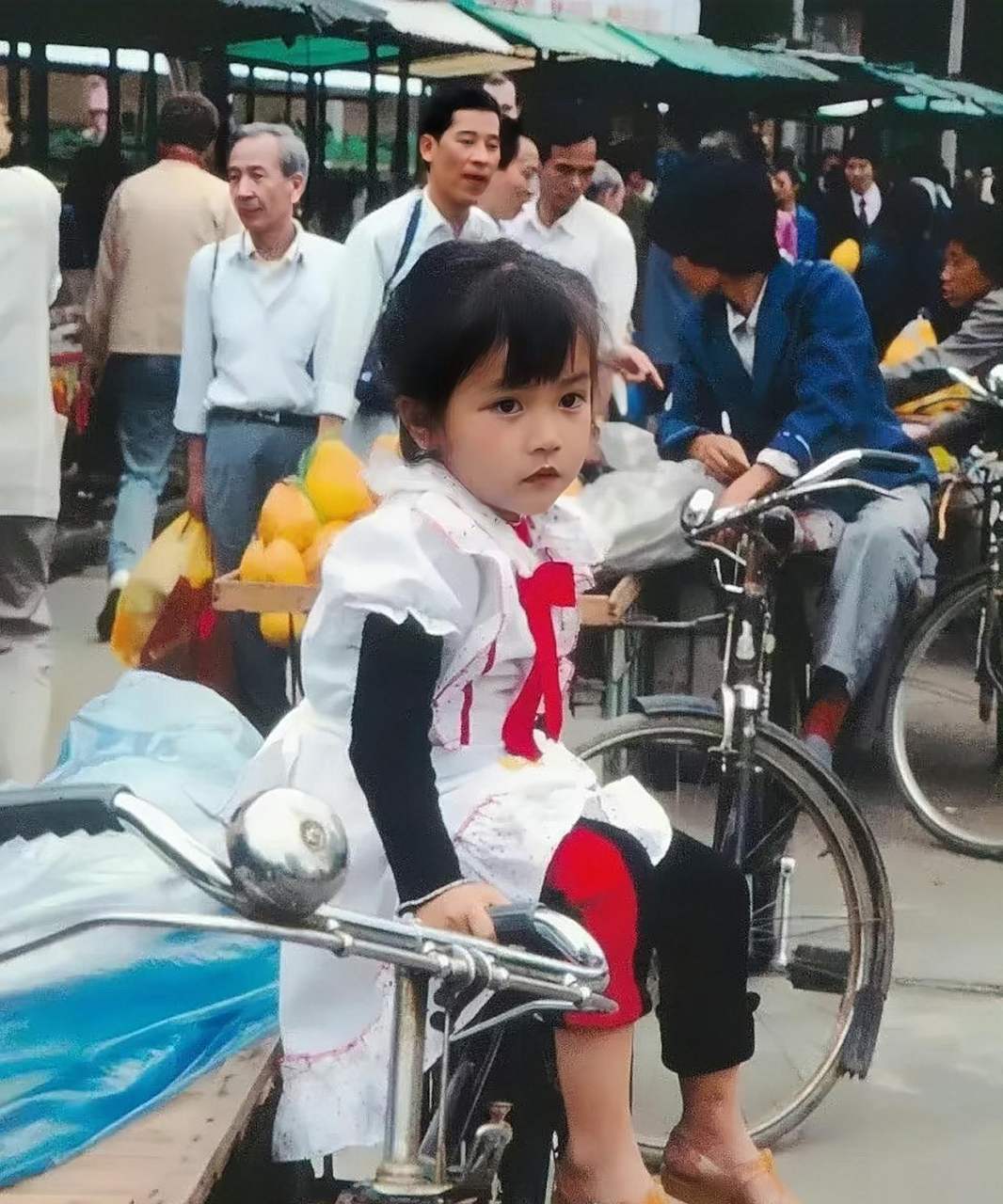 一个大眼睛的小姑娘乖乖的坐在一辆凤凰自行车的后座上,等着在市场里