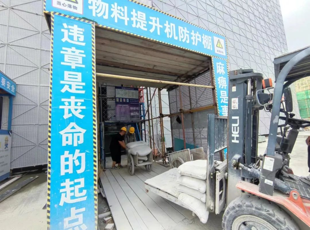 一名司机开叉车将水泥运到物料提升机防护棚入口,两位工人把水泥搬到