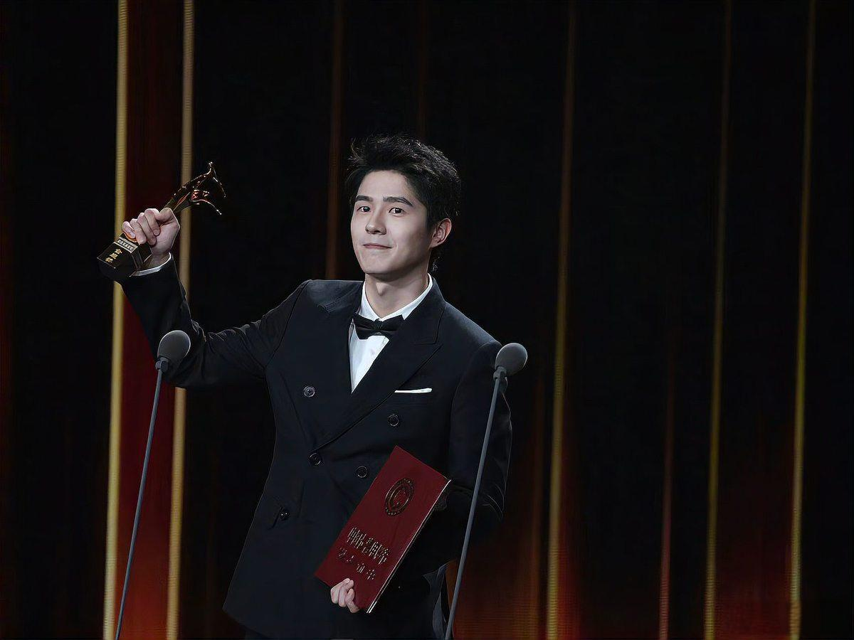 刘昊然青年男士在颁奖典礼上手持奖杯和证书,身穿黑色西装,背景为舞台