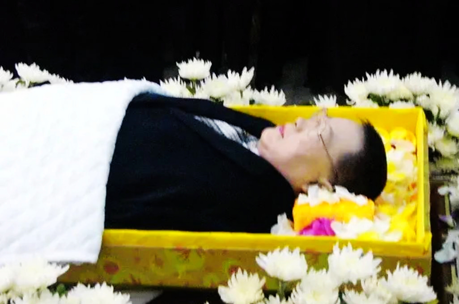 照片中,他戴着眼镜,身着黑色西装,安详地躺在花丛中.